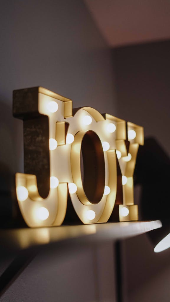 Close-up of a lighted 'Joy' sign with warm bulbs on a shelf, indoors.